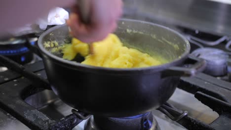 Closeup-of-female-cook-stirring-corn-meal-with-wooden-spoon-in-black-pot-on-gas-stove,-woman-preparing-traditional-polenta-in-kitchen