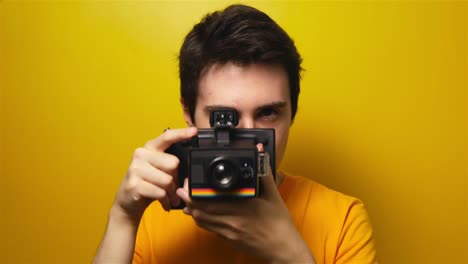 Portrait-of-attractive-young-millenial-man-taking-pictures-on-vintage-film-camera-on-yellow-background