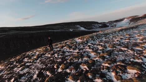 Un-Hombre-Parado-En-El-Borde-De-Una-Montaña-Erosionada-Con-Hielo-Derritiéndose-Y-Un-Gran-Paisaje-De-Una-Puesta-De-Sol-Ardiente-En-Islandia