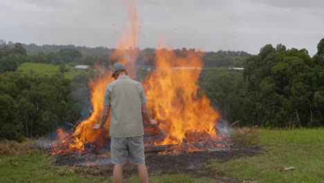 A-man-in-a-beanie-stands-in-front-of-a-raging-bonfire-on-a-bush-property