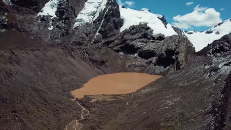 Aerial,-reverse,-drone-shot-away-from-a-brown-water-lake-underneath-snowy-mountain-peaks,-in-Cusco-region,-Andes,-Peru