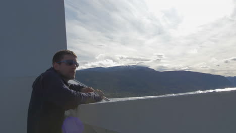 A-Man-in-Sunglasses-Looks-out-over-the-Edge-of-a-Ferry-Taking-in-a-Nice-View-of-Mountains-on-a-Sunny-Day,-Slow-Motion