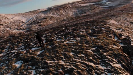 Ein-Mann-In-Einer-Vr-brille,-Der-über-Der-Steilen-Hochlandklippe-Steht,-Mit-Dünnen-Schneeschmelzen-Auf-Felsrissen-In-Island---Drohnenrotationsaufnahme