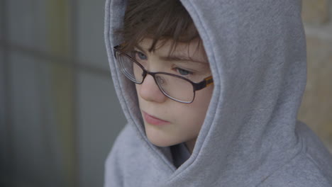 Cute-boy-in-a-gray-hoodie-and-glasses-waiting-outside-looks-around,-closeup
