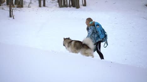 Young-woman-with-backpack-walks-with-big-dog,-snowy-winter-scene,-slowmo
