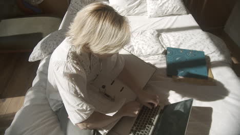 Adult-woman-works-from-home-on-bed-with-laptop-computer,-high-angle-pan