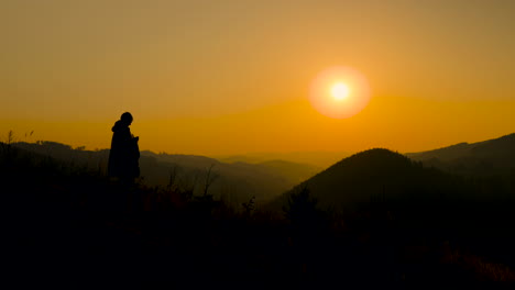 The-silhouette-of-a-woman-watching-the-sunset-and-sitting-down-stump-looking-out-at-sunset-with-dark-colors-of-mountains-and-hills-in-the-background-along-with-a-orange-sun-in-the-background-4k-60fps
