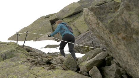 A-Woman-Hiking-Up-Steep,-Rocky-and-Snowy-Terrain-Using-a-Rope-on-an-Overcast-Day-in-Norway,-Slow-Motion