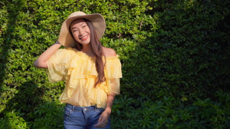 Static-shot-of-a-beautiful-asian-lady-wearing-a-summer-hat,-yellow-top-and-jeans-turning-around-while-smiling-at-the-camera