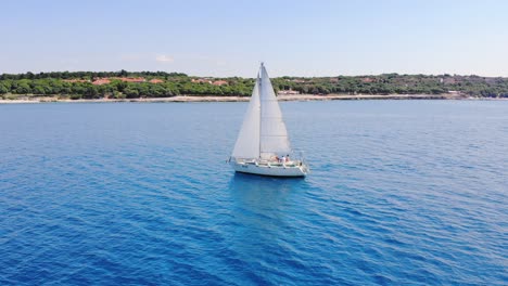 Ein-Einsames-Weißes-Segelboot-Segelt-Entlang-Der-Grünen-Küste-Unter-Wolkenlosem-Himmel-Auf-Ruhigem,-Klarem,-Blauem-Wasser