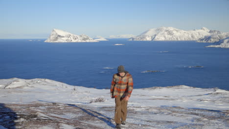 A-Man-Hiking-Up-a-Snowy-Landscape-with-the-Arctic-Ocean-and-Mountains-in-the-Background,-Slow-Motion