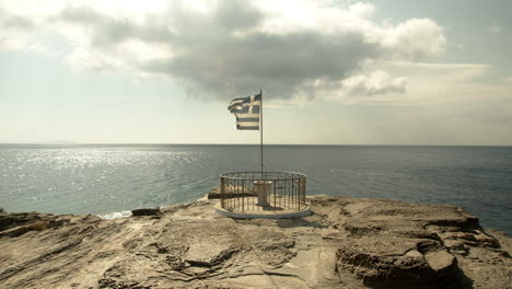 4k-wide-shot-of-a-Greek-flag-waving-in-slow-motion-with-the-sea-in-the-background,-on-a-rocky-shore
