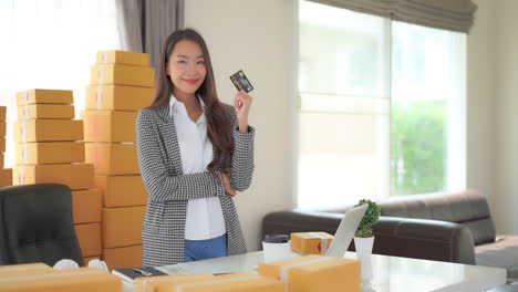 Static-shot-of-a-cute-Asian-lady-standing-in-an-office-surrounded-by-boxes-while-holding-a-credit-card-in-one-hand-and-doing-a-thumbs-up-in-the-other-hand
