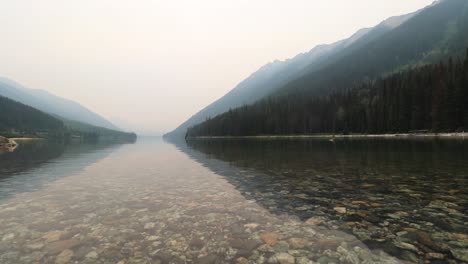 Low-angle-wide-shot-at-a-lake,-transparent-water-visible-stones-at-the-bottom,-reflection-of-mountains-and-forest