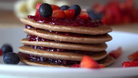 Blueberries-falling-down-on-top-of-stacked-pancakes-with-red-jam-and-fresh-strawberries,-SLOW-MOTION