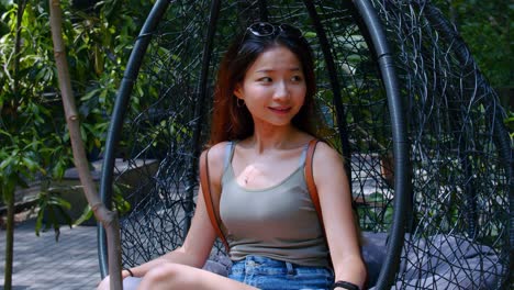 Joven-Asiática-Sentada-En-Una-Silla-Colgante-Sonriendo-Mientras-Mira-Alrededor-Del-Parque-Brisbane,-Australia