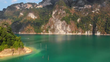 Viridisches-Wasser-Des-Cheow-Lan-Dam-Mit-Wunderschönen-Bergen-Im-Hintergrund-Und-Zwei-Longtail-booten,-Die-In-Der-Ferne-Auf-Dem-See-Segeln,-Thailand