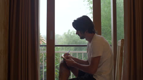 Man-Writing-and-Staring-Out-of-Window-in-Isolation-on-Rainy-Day