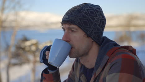 A-Man-in-a-Snow-Hat-Calmly-Sips-Tea-out-of-a-Mug-with-a-Snowy-Backdrop,-Slow-Motion