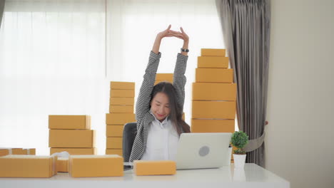 A-young-Asian-woman-surrounded-by-products-to-be-shipped-stretches-after-working-long-hours-on-her-laptop