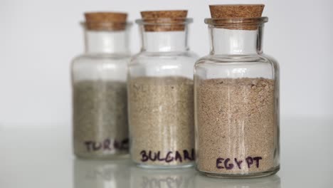 Hand-of-caucasian-male-placing-small-transparent-jars-filled-with-sand-from-different-countries-next-to-each-other-on-white-background
