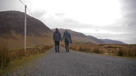 Couple-holds-hands-while-walking-road-in-Connemara,-low-angle-rear-view