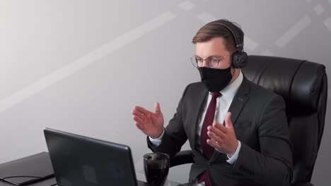 Businessman-working-in-home-office-in-protective-mask-communicating-on-webcam-in-headphones