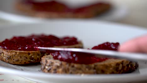 Male-hand-spreading-sweet-red-strawberry-jam-on-top-of-golden-brown-toasted-flax-seed-bread-with-silver-knife
