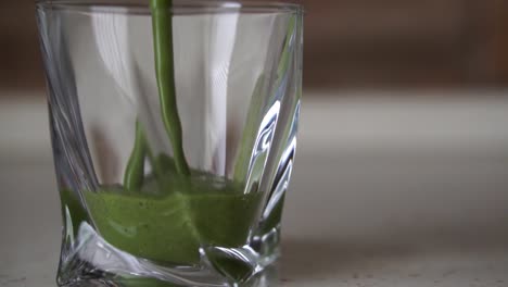 Pouring-thick-green-smoothie-into-twisted-crystal-glass-sitting-on-countertop,-SLOW-MOTION