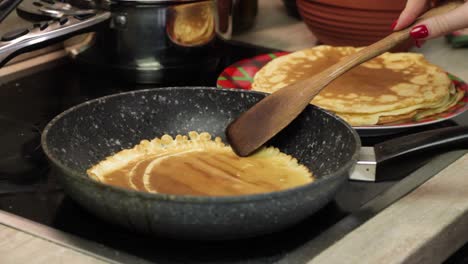 Caucasian-woman-hand-with-red-nail-polish-turns-over-pancake-in-black-skillet-on-stove