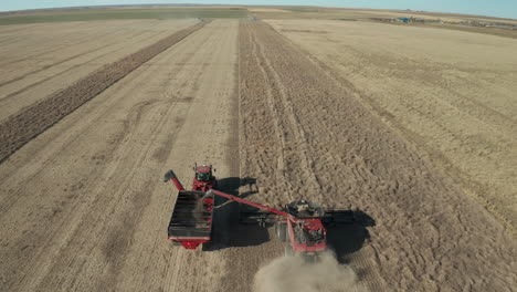 Große-Industrielle-Landwirtschaftsmaschine-Erntet-Weizenkorn-Und-überträgt-Ernte-In-Einen-Roten-Lkw-In-Einem-Flachen,-Weitläufigen-Ackerland-In-Der-Landschaft-Von-Ponteix,-Saskatchewan,-Kanada,-überkopfanflug