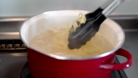 Cooking-Pasta-In-A-Red-Pot-With-Boiling-Water-Using-Tongs---Closeup-Shot
