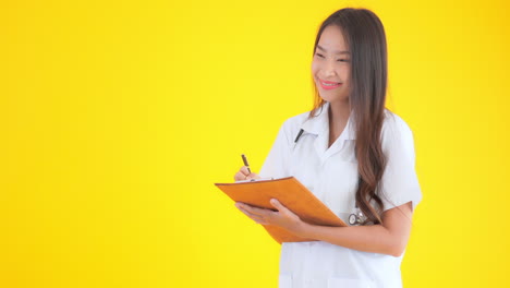 Asian-nurse-doctor-woman-writing-notes-in-the-clipboard-on-yellow-isolated-background-smiling