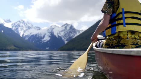 Young-Man-With-Yellow-Life-jacket-In-Calm-Lake-Rowing-Towards-Snowy-Mountains