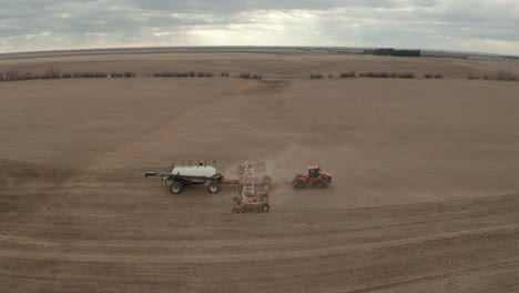 Panoramic-sideways-aerial-view-above-red-industrial-seeding-machine-pulling-fertilizer-in-flat-farm-field-in-rural-countryside-on-cloudy-day,-Swift-Current,-Saskatchewan,-Canada