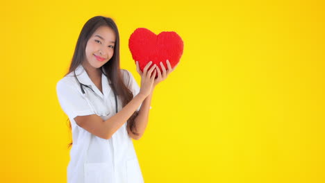 Asian-woman-nurse-wearing-white-uniform-and-stereoscope-showing-fluffy-red-heart-cushion-smiling