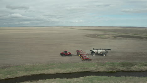 Aerial-scenic-view-of-red-industrial-tractor-seeding-machine-pulling-fertilizer-in-flat-farm-field-in-expansive-rural-countryside-on-cloudy-day,-Vanguard,-Saskatchewan,-Canada