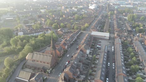Drohnenblick-Hinab-Zu-Britischen-Stadthäusern---Kirche-Mit-Kirchturm-Bei-Sonnenaufgang,-Während-Autos-Eine-Lange-Straße-Hinunterfahren