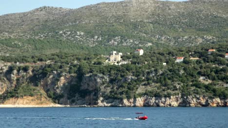 Blick-Auf-Hügel-Und-Klippen-In-Kroatien-über-Meerwasser-Mit-Kleinem-Roten-Boot,-Das-Vorbeisegelt