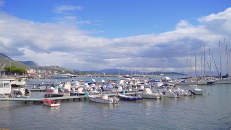 Scenic-view-of-white-motorboats-and-sailboats-tethered-to-floating-docks-at-Gaeta-marina-sea-port-with-mountain-range-in-background-on-sunny-day,-Italy,-static