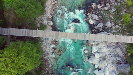 Drone-spirals-down-toward-footbridge,-wild-mountain-river-flows-below