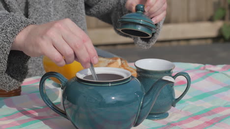Hand-takes-lid-from-teapot-to-stir-at-outdoor-breakfast-table,-slow-motion