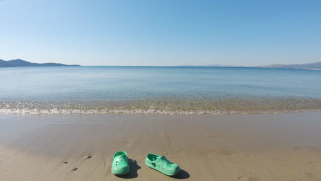 Green-Crocs-pulled-out-by-waves-on-sand-beach-on-sunny-day,-slomo