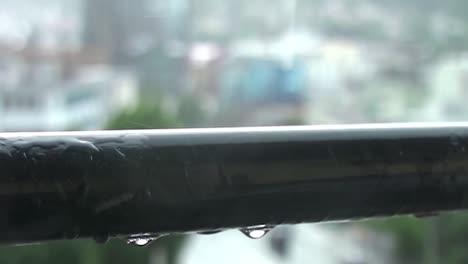 Close-up-of-rain-drops-falling-off-a-horizontal-metal-rod-on-a-balcony-with-houses-and-trees-in-the-blurred-background