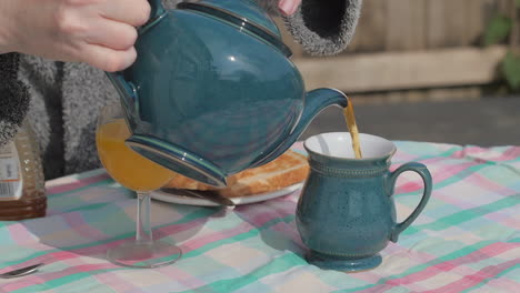 Person-pours-tea-from-pot-into-mug-on-outdoor-breakfast-table,-medium-shot