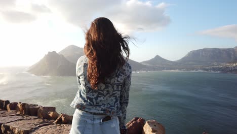 A-beautiful-bay-surrounded-by-mountains-with-a-long-dark-haired-beautiful-girl-looking-out-towards-the-waters-as-the-camera-circles-around-her-in-an-arc