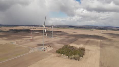 Hohe-Windkraftanlagen-Ragen-über-Braune-Landwirtschaftliche-Felder-Mit-Gewitterwolken-Und-Regen-In-Der-Ferne