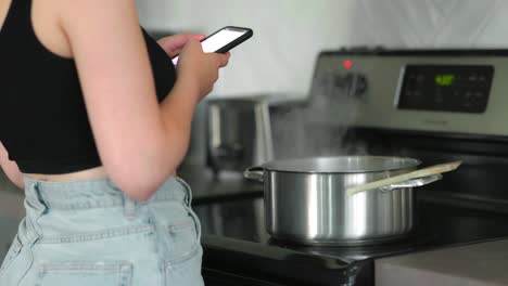 Caucasian-woman-standing-in-front-of-stove,-on-her-phone,-as-steam-rises-from-the-pot-of-cooking-pasta-on-the-stove