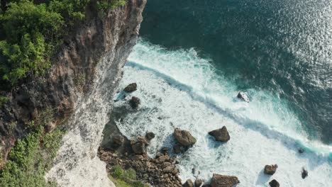 Drone-flying-over-tall-seaside-cliff-revealing-rocky-beach-with-dark-blue-water-and-waves-crashing-into-the-shore