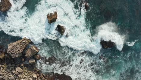 Aerial-top-down-view-of-ocean-waves-crashing-into-rocky-shore-with-large-rocks